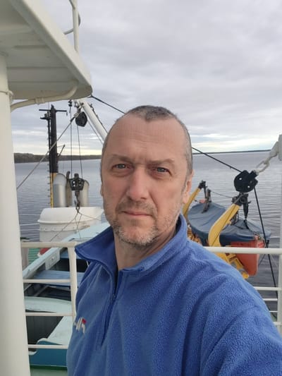 A medium shot depicts a middle-aged man with short, graying hair and a trimmed beard, wearing a blue fleece jacket. The man is the primary subject and is positioned off-center to the right. He appears to be taking a selfie.
The background shows elements indicating a boat or ship environment. Visible are white railings, metal equipment including davits and lifeboats, and a body of water. Across the water is a distant shoreline with sparse vegetation.
The sky is overcast with dull, gray clouds. Based on the positioning and background details, the photograph could be taken on a vessel on a river or lake near a shoreline. The location mentioned in the instructions is Shcheleiki, Russia, but determining this from the image alone is not possible without additional details.