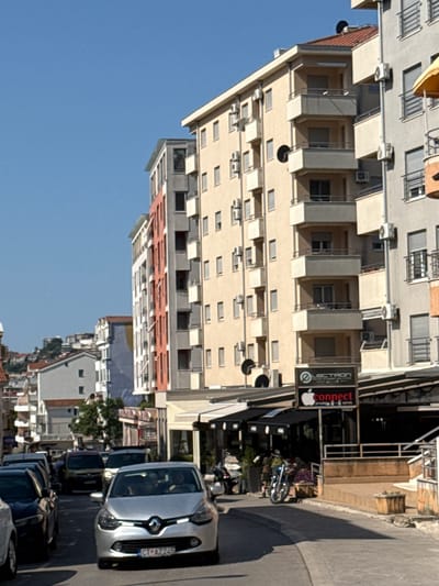 The image captures a street scene in Lapčići, Montenegro, under a clear blue sky. The perspective is from within a vehicle, looking forward down a paved street lined with multi-story buildings. On the right side of the street, a modern building features balconies with metal railings and external air conditioning units. A sign with the text "LECTRON" and a red apple logo with "connect" below it is visible on the ground floor of this building. To the left, another building has a facade with alternating sections of white and coral-colored panels. Further down the street, more buildings and some greenery are visible. Traffic is present, with several cars in front, including a silver Renault Clio in the immediate foreground with license plate "CT AZ 245". A motorcycle is parked on the right side of the street. The overall impression is of a sunny day in a populated urban area.