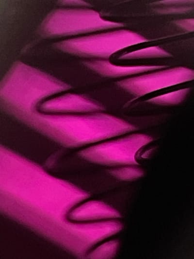 The image displays an abstract, close-up view characterized by a repeating pattern of dark, curvilinear forms set against a vibrant magenta background. Multiple parallel, wave-like or zigzag dark lines, consistent with shadows or three-dimensional coiled/bent objects, traverse the frame. These dark elements contrast sharply with the illuminated magenta surfaces, which show minor variations in brightness and a slightly grainy texture. The right side and bottom left portions of the image are dominated by solid black areas, indicating deep shadow or an unlit environment. No discernible people, specific objects beyond the abstract forms, actions, or interactions are present. The precise location in the city of Derby, United Kingdom, cannot be determined from the visual information provided.