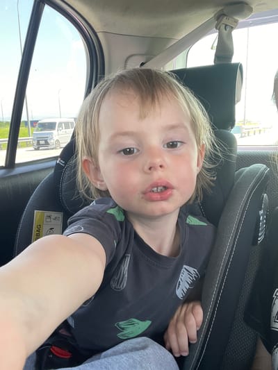 The image shows a young child with blonde hair and brown eyes sitting in a car seat, facing the camera. The child is wearing a dark grey t-shirt with a green pattern. The car seat is black with grey stitching and has an airbag warning label on the side. In the background, through the car window, a white van is visible on a road with streetlights and vegetation. The environment appears to be outdoors, possibly on the outskirts of a city, under a bright sky. The location is identified as Solovej-Kljuch, Russia.