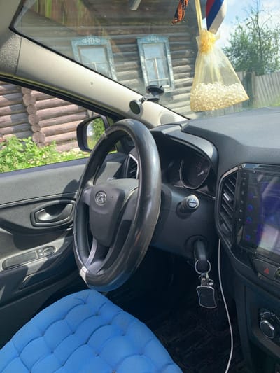 This media file shows the interior of a car parked outside a wooden house in what appears to be a rural setting in Barsai, Russia. The sunlight suggests it is daytime, possibly morning or afternoon.
The primary subjects are the car's interior elements: a steering wheel, dashboard with a touch screen display, car keys in the ignition, and a portion of a blue upholstered seat. Hanging from the rearview mirror or similar attachment point on the windshield is a decorative item consisting of a ribbon with red, white, and blue stripes and a small, sheer golden pouch filled with what looks like small, pearl-like beads. A dark fabric ribbon with an orange and black pattern is also visible, possibly a St. George's ribbon, often associated with patriotic or remembrance events.
The scene is viewed from the perspective of someone sitting inside the car, looking towards the dashboard and passenger side. Through the windshield, a rustic wooden house with two windows featuring decorative white trim is visible. The exterior appears to be a sunny day with some greenery and trees in the background, suggesting a natural, possibly summer environment.
There is no visible text in the image, except for what might be on the car's display screen, which is too blurry to decipher. The overall impression is a snapshot of a car interior on a bright day, with elements that hint at a cultural context within Russia. No people are visible, and no specific activity is taking place other than the car being parked.