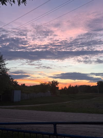 The image captures a rural landscape at dusk. The sky is filled with dramatic clouds displaying hues of purple, pink, and orange, indicating either sunrise or sunset. Several power lines stretch horizontally across the upper portion of the frame. In the midground, a line of trees forms a dark silhouette against the colorful sky. In front of the trees, a weathered wooden fence encloses an area with a small, light-colored shed. A dirt road or path is visible in the foreground, leading towards the treeline. The overall impression is of a serene, natural setting in a rural area, likely a village or countryside. The specific location of Ганчарскі сельскі Савет, Belarus, places this scene in a particular administrative region of Belarus, known for its rural character.