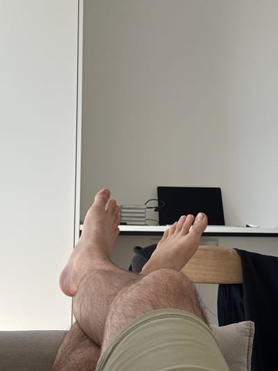 The image, when correctly oriented, captures a close-up view of a person's legs and feet in an indoor setting. The majority of the background consists of a plain, light-colored wall. On the right side of the background, a dark, rectangular screen or monitor is mounted on the wall, with cables visible beneath it.

In the foreground, a person's legs are prominently featured. The right leg, which is hairy, is bent at the knee, with the lower leg and foot extended towards the left. The foot is bare, showing toenails. A part of the person's left foot is also visible further back in the frame. To the immediate right of the person's legs, a section of a grey, upholstered sofa or chair with a matching cushion is visible. A light-colored wooden surface, possibly a desk or shelf, is situated behind the legs, with a dark garment draped over it.

The lighting is bright and even, indicating it is daytime. The overall ambiance suggests a casual and relaxed atmosphere, with the person likely lounging or resting. There are no specific activities depicted beyond this state of repose. No visible text is present in the image. The setting appears to be a modern interior space, consistent with a residence or hotel room in Kitsjanovo, Russia.