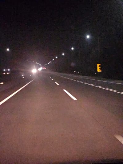 The image captures a nighttime view of a highway, likely in Sangju-si, South Korea, characterized by a dark sky illuminated by streetlights and vehicle headlights. The road, paved with asphalt, has distinct white lane markings, a solid white line on the left and dashed white lines indicating traffic lanes. Several vehicles are visible: on the left, headlights suggest a vehicle approaching, while further ahead, a line of headlights indicates a procession of cars. On the right side of the highway, a yellow rectangular sign with black silhouettes of deer is illuminated, warning of wildlife crossing. The overall impression is a well-lit highway at night, with traffic moving, and a reminder of the potential presence of animals.