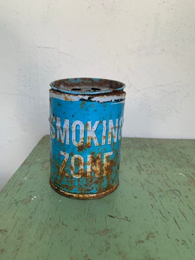 The image shows a close-up of an old, rusted blue metal ashtray with "SMOKING ZONE" printed on it in white letters. The ashtray is placed on a distressed green wooden surface. The background is a textured white wall. There are no people visible in the image. The scene appears to be indoors or in a covered outdoor area. The location in Silmala, Latvia, is not determinable from the visual information provided.