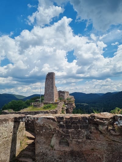 The image shows the ruins of a castle on a hill overlooking a valley in Dahn, Germany. The castle ruins are made of stone and appear to be ancient. There is a tall, broken tower in the center of the ruins, and other stone walls and structures are visible. The surrounding landscape is hilly and covered in trees. The sky is blue with white clouds. There are no people in the image.