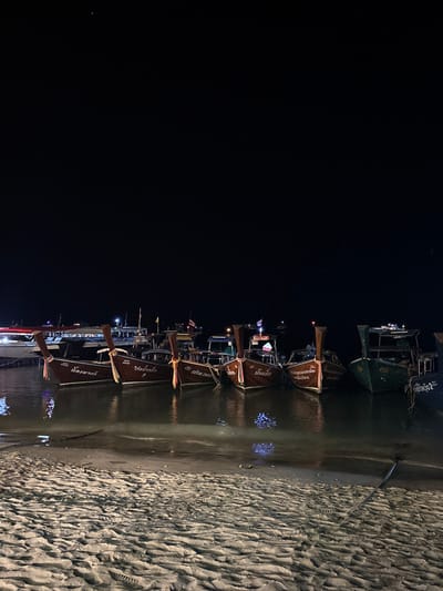 The image depicts a nighttime scene along the shoreline of Ban Ko Li Pe, Thailand. In the foreground, light-colored sand exhibits distinct ripple patterns and faint indentations, indicating a wet surface. The midground is dominated by a fleet of approximately eight boats moored in calm water near the shore. Prominently featured are at least five traditional Thai longtail boats, constructed from dark wood, displaying high pointed bows and tall stern posts. These boats bear Thai script on their hulls, likely names or identifiers, and are equipped with small flags (including the Thai national flag) and low-level artificial lights that reflect on the water's surface. A larger, lighter-colored vessel is partially visible on the far left, and a boat with a green hull is situated towards the right of the longtail fleet. The background is a dark, clear night sky, with minimal distant artificial light sources visible above the horizon. No human figures are present.