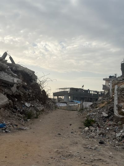 The image depicts a devastated urban landscape in Jabalia, Palestinian Territory, characterized by extensive structural damage and debris. A central, unpaved dirt path, approximately 2-3 meters wide, extends into the distance, narrowing slightly. On both sides of this path are large mounds of rubble, composed of broken concrete, twisted rebar, masonry fragments, and other building materials, indicating collapsed structures.
To the left, a substantial pile of debris forms a steep slope, showing exposed rebar protruding upwards and outwards. To the right, similar rubble piles line the path, with some standing, fractured wall sections visible further back. Small patches of sparse vegetation are discernible among the debris.
In the mid-distance, several buildings appear heavily damaged or partially destroyed. One building on the left horizon features an exposed framework, while another structure further along the path shows missing sections and exposed interiors. A dark, possibly partially collapsed, multi-story building is visible in the center-right background, with a small satellite dish mounted on its upper level. Minimal, makeshift structures, possibly constructed from corrugated metal, are visible near the distant buildings. The sky above is overcast with scattered clouds, and no individuals or active interactions are present in the scene.