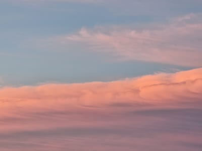 The image depicts a sky filled with clouds displaying hues of pink, orange, and pale blue, suggestive of a sunset or sunrise. The clouds are layered, with a prominent band of vibrant pink and orange clouds occupying the middle and lower portions of the frame. Finer, wispy clouds with a lighter pinkish tint are visible in the upper part of the sky, against a backdrop of pale blue. The scene appears to be an outdoor, natural environment, likely an open sky view. No people, specific landmarks, or structures are visible. The image is a photograph of the sky.