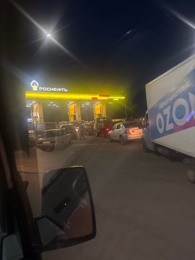 The image depicts a nighttime scene at a gas station, likely in Petropavlovka, Russia, given the context. The primary focus is a queue of vehicles lined up at the pumps under bright yellow illumination from the gas station's canopy. The "РОСНЕФТЬ" logo, indicating Rosneft, a Russian oil company, is prominently displayed on the canopy.

Several cars are visible in the queue, and a large truck with "OZON" branding on its trailer is also part of the scene, positioned at the front of the line. The weather appears to be dark, typical of nighttime, with no strong indicators of precipitation.

A side mirror of a vehicle is in the foreground on the right, partially obscuring the view of the gas station and the cars. License plates are visible on some of the vehicles, though the text is difficult to decipher clearly. One visible license plate appears to be "W246KP 34".

The overall impression is one of a busy gas station during nighttime hours, with vehicles waiting to refuel. The bright lights of the station stand out against the dark background, creating a functional and somewhat stark atmosphere. No individuals are clearly discernible in the image.