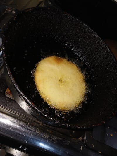 A close-up, high-angle shot shows a circular, golden-brown food item being deep-fried in a dark, cast-iron pan. The food item is submerged in hot oil, which is bubbling vigorously around its edges. The pan itself appears well-used, with some residue visible on its surface. The pan rests on what appears to be a gas stove, indicated by the metallic grates and burners visible at the bottom of the frame. The lighting is dim, with the primary light source appearing to come from above, illuminating the frying food. The background is dark and indistinct.
