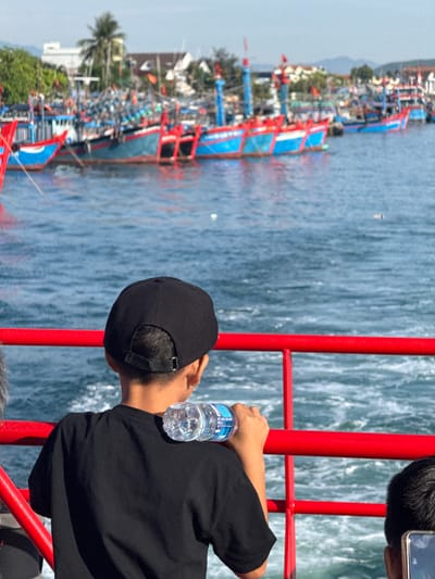 The image depicts a young boy wearing a black t-shirt and a black baseball cap, seen from behind as he stands on a boat. He is holding a clear plastic bottle of water, with the bottle resting on his shoulder. In the background, a fleet of brightly colored fishing boats, predominantly blue and red, are docked in the calm, blue water. Palm trees and buildings are visible on the shoreline, suggesting a tropical coastal setting. The scene is likely located in the city of An Hải, Vietnam, a known area with a significant fishing industry and harbor. The bright sunlight casts a clear illumination on the scene, highlighting the vibrant colors of the boats and the texture of the water.