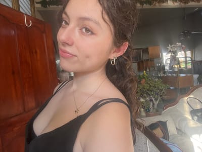 A woman with curly brown hair poses for a selfie. She wears a black top with thin straps and a gold necklace. She has an ear piercing and a nose ring. In the background, there's a wooden door, a mirror reflecting a room with chairs, cabinets, and a plant decorated with purple ornaments. Part of a table with luggage on it is also visible.
