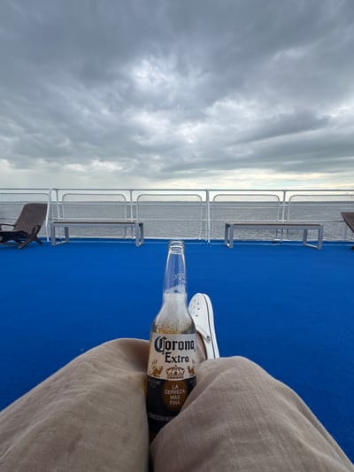 The image shows a close-up view of a person's legs and a bottle of Corona Extra beer on a bright blue deck, likely on a ferry or cruise ship. The person is wearing light-colored pants and white footwear. In the background, there is a railing, benches, and chairs, all facing the water. The sky is overcast with gray clouds. The scene is not located in Belous, Russia, as no identifiable landmarks or context suggest this location.
