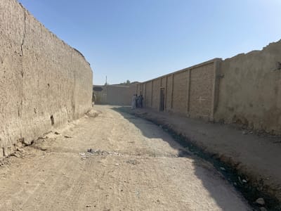 This image depicts an unpaved, dusty thoroughfare in Spin Boldak, Afghanistan, under a clear, bright blue sky. The scene is dominated by earth-toned architectural structures and natural elements.
On the left side, a tall, aged mud-brick wall extends from the foreground into the midground, exhibiting vertical cracks and erosion at its base. A small, dark, roughly rectangular aperture is visible in this wall approximately one-third of the way from the foreground. The wall gradually curves to the right in the midground.
On the right side, a shallow, earthen ditch containing sparse green vegetation and debris runs parallel to the path. Beyond this ditch, a long wall is present. The lower section appears to be a light-colored render or mud-brick, while the upper section consists of exposed brown bricks with vertical pilasters at regular intervals. A dark, rectangular wooden door is set into this brick section. Further down, this wall transitions back to an irregular, weathered mud-brick construction.
The central dirt path shows visible tire tracks and scattered debris. In the immediate foreground, a small pile of dark, charred material or ash is present on the path.
Approximately three to four adult male figures, dressed in long garments, are visible in the midground, standing near the brick wall on the right. Their specific actions are not discernible due to distance. Another partially obscured figure is visible further down the left wall.
In the distant background, additional low-rise, earth-colored structures are visible, along with a faint, slender pole-like object protruding against the horizon. The overall impression is one of a dry, rural, or semi-urban environment constructed primarily from local earth materials.