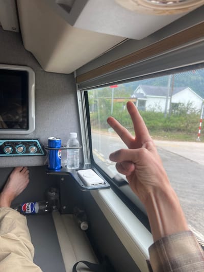 The image depicts the interior of a passenger vehicle, likely a sleeper bus, looking outwards through a window onto a roadside environment in M'Drắk, Vietnam.
**Interior:**
On the left, a bare human left foot is visible, resting on a beige upholstered seat or footrest. Further up and left, a small, dark-screened monitor with a white bezel is embedded in a grey fabric wall. Below the monitor, a control panel features two circular dials (one blue, one silver) and multiple smaller buttons, with partially visible text including "Light" and "USB." A black metal console extends from the wall towards the window, featuring two circular cup holders. The left holder contains a blue Pepsi soda can; the right holder contains a clear plastic bottle of water. To the right of the cup holders, on an integrated flat tray, rests a white portable power bank with a white circular pop socket attached. Below the console, on the vehicle floor, two empty plastic Pepsi bottles are present. A black strap, possibly from a bag, is also visible. The vehicle interior features grey fabric wall coverings and beige/off-white seating and trim.
**Human Element:**
In the right foreground, a pale-skinned human right arm, wearing a sleeve of a brown and white plaid shirt, is extended towards the window. The hand is performing a "peace" or "V for victory" gesture with the index and middle fingers extended upwards, while the ring and pinky fingers are curled into the palm. Visible veins are present on the forearm.
**Exterior Environment (M'Drắk, Vietnam):**
Beyond the window, an asphalt road with a yellow double line marking is visible. The immediate roadside features green grass and vegetation. In the mid-ground, a white gabled house with a dark roof stands amidst greenery, alongside a smaller, partially obscured structure. A red rectangular sign or marker is visible near the house, and a white and red striped marker pole stands to the far right. The background consists of lush green hills or mountains, suggesting a rural or semi-rural setting. The lighting appears bright and overcast.