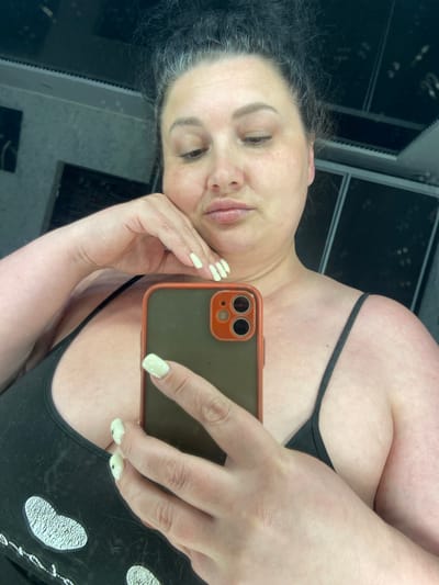 A woman with dark, curly hair pulled up in a bun and light-colored nail polish on her nails is taking a selfie in a room. She is wearing a black tank top with a white graphic design on the front. Her left hand is resting on her chin. She is holding a smartphone with an orange and green case in her right hand to take the photo. The background appears to be a room with reflective surfaces and ceiling lights, suggesting it might be a dressing room or a room with mirrors. There is no visual information available to determine the location in the city of Rozkvit, Ukraine.