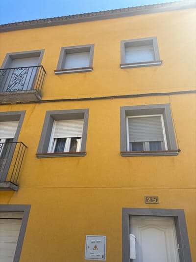 The image shows the exterior of a yellow building in Mallén, Spain. The facade features several square windows with gray frames. Some windows have blinds or security grilles. Above one of the windows, the number "25" is displayed. A white electrical box with a lightning bolt symbol is visible near the upper right corner. Electrical wiring runs vertically along the building's surface. A glimpse of the blue sky is visible at the top left corner.
