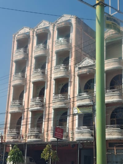The image shows a peach-colored, multi-story building with ornate balconies and arched windows, located on a street in Bireuenmeunasahcapa, Indonesia. A tall, green utility pole with a yellow sign displaying "DISEWAKAN" and a phone number is positioned in the foreground, partially obscuring the building. Overhead power lines crisscross the scene. Below the building's balconies, there are storefronts, including one with a sign for "MAILA OPTIK". A white van is visible parked in front of one of the storefronts. The sky is clear and blue. The building appears to be a commercial or residential property for rent.
