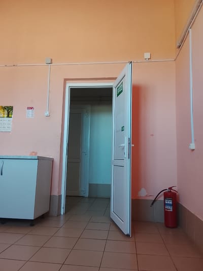 An interior scene depicts a brightly lit room with peach-colored walls and beige tiled floors. A partially opened white double-door with a glass panel sits in the center of the frame. The door on the right is wider and opens inwards, revealing a glimpse of a hallway with a similar tiled floor and beige walls. A green sign with white Cyrillic lettering that reads "ВХОД" (meaning "Entrance") is affixed to the glass panel of the open door.
To the left of the doors, a white cabinet with a silver handle stands against the wall. A calendar with a nature scene and some printed text is attached to the wall above the cabinet, accompanied by a red and white sign displaying Cyrillic text and numbers.
On the right side of the image, near the corner, a red fire extinguisher rests on the floor against the peach-colored wall. A white electrical conduit runs along the wall, terminating in a white electrical box with a single outlet. Another similar electrical box is visible higher up on the wall near the ceiling. The overall environment appears to be an institutional setting, possibly a school, clinic, or office building. There are no people visible in the image. The image quality suggests it was taken with a mobile phone camera.