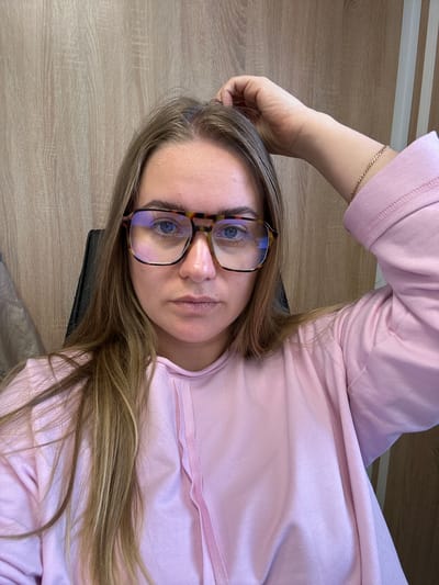 A woman with light brown hair and blue eyes wearing tortoiseshell glasses and a pink shirt is lying down. She is wearing a gold bracelet on her left wrist. The background appears to be a light-colored wooden surface. The location in the city of Ural'skoe, Russia, is not discernible from the image.