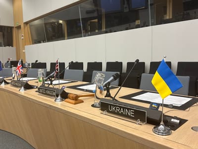 The image depicts a conference table in a room with a large glass window. The table is covered with a light wood surface and there are several microphones placed in front of nameplates that read "United Kingdom" and "Ukraine". There are several flags on the table, including the Ukrainian flag, the British flag, and a flag of the European Union. There are also microphones and glasses of water on the table. The room is located in Brussels, Belgium, at the coordinates 50.879, 4.422. The background includes several empty chairs, and a large glass window with a view of an office environment. There are also two glass panels on the wall to the left of the room. There is a wooden gavel on the table. There are no people visible in the image.