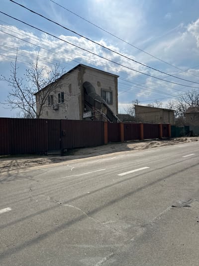 The image shows a residential street in Horenka, Ukraine (latitude: 50.525, longitude: 30.241) with a two-story house on the left side of the street. The house is damaged and appears to be in the process of being repaired, as scaffolding is visible. There is a brown fence in front of the house, and a second house is visible in the background. The street is paved with asphalt and has white markings for traffic lanes. The sky is partly cloudy with some sunlight shining through. The scene is empty of people.
