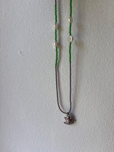 This is a horizontal, eye-level, medium shot of a necklace lying on a textured white surface. The necklace is primarily composed of small, dark gray beads strung together, forming a loop. Interspersed along the top strand of the necklace are three flat, oval, white beads. The bottom strand of the necklace also has three similar white beads. Attached to the dark gray beaded strand on the right side of the image is a small, silver-colored charm shaped like a frog. The frog charm has small red accents on its eyes. The background is a textured, slightly uneven white surface, possibly a wall, with subtle shadows suggesting overhead lighting. No people, text, or specific events are visible. The setting appears to be indoors.