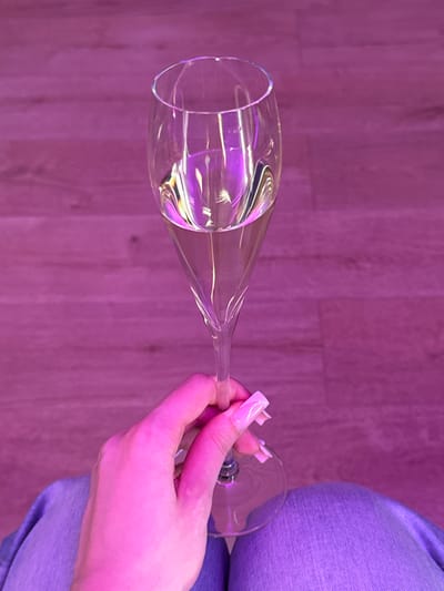 An overhead shot shows a person's left hand holding a champagne flute filled with a pale, clear liquid, lit by a pink light that casts purple and pink reflections on the glass and the wooden floor beneath. The hand has long, square, pale pink fingernails. The person is wearing blue denim jeans. The environment appears to be indoors with wooden plank flooring. There is no visible indication of the location being the city of Zakharovo, Russia.