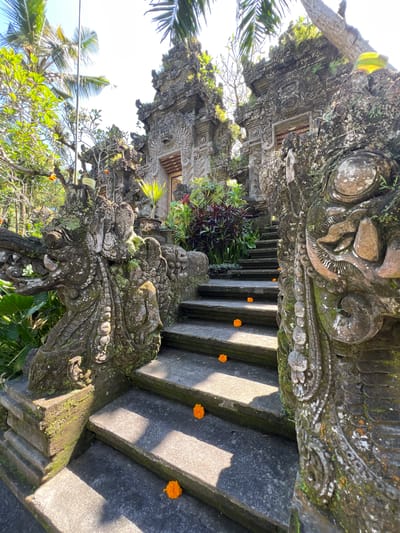 This photograph shows a set of stone steps in Kedewatan, Indonesia. The steps are adorned with intricately carved stone statues covered in moss and vegetation. Small orange flowers are scattered on the steps. Lush greenery is visible around the steps and stone structures with carvings. The scene appears to be a pathway leading toward the entrance of a temple.