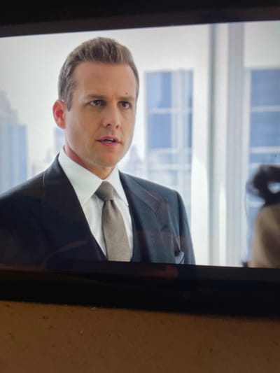 The image displays a close-up of a Caucasian male from the chest up, seemingly captured from a screen. The man has short, styled brown hair, a clean-shaven face, and his mouth is slightly ajar as if speaking. He wears a white dress shirt, a light-colored tie (beige/taupe), and a dark suit jacket over a waistcoat, with a dark vest visible beneath. The background shows a brightly lit, modern office space with large windows revealing a blurred urban cityscape featuring tall buildings. A dark, indistinct shape, possibly the back of a head or office furniture, is visible in the far right background. The scene is located in the city of New Forest, United Kingdom.