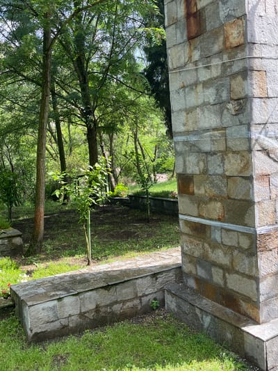 The image displays a green, arboreal environment in Aytos, Bulgaria, partially framed by a substantial stone structure on the right and bottom. A vertical stone pillar, composed of rectangular blocks of varying grey, brown, and reddish-brown hues, occupies the right side of the frame. Two horizontal metallic or plastic strips are visible on the pillar at different heights. At the base of the pillar, a lower stone structure or bench extends towards the left, connecting with a longer, low stone wall or planter in the bottom-left quadrant. Both lower stone elements appear damp. The ground is covered with green grass interspersed with areas of bare soil. In the midground, several young saplings with bright green leaves are visible, one of which is supported by a slender stake. Behind these, numerous mature deciduous trees with dense green foliage dominate the left and center-left background, creating a thick canopy through which bright daylight filters. No individuals or animals are present.