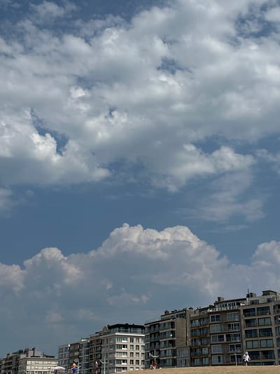 The image shows a view of the coastline in Koksijde, Belgium, with a clear blue sky filled with dramatic cumulus clouds. In the foreground, several multi-story apartment buildings line the beach, their facades characterized by numerous windows and balconies. The buildings appear to be modern and residential, with a mix of lighter and darker architectural elements. Below the buildings, a sandy beach is visible, with a few people scattered about. One person is sitting on the sand, while another is standing near the buildings. Further down the beach, a person wearing a white hat and shirt is walking away from the camera. The overall scene captures a pleasant day at the seaside, with the imposing cloud formations adding a sense of depth and scale to the urban coastal landscape.