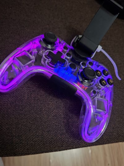 The image displays a transparent video game controller with purple and blue internal lighting, resting on a dark brown, textured surface, likely a couch or upholstered chair. A black phone mount is attached to the top of the controller, and a white cable extends from the controller's base. The controller's various buttons, joysticks, and transparent casing are visible, with the lighting creating a vibrant glow. There are no people visible in the image. The scene is indoors. There is no information within the image that can be used to determine its location in Ilava, Slovakia.