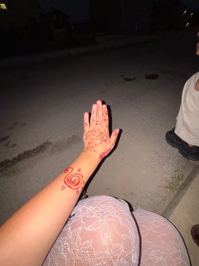 The image shows a person's arm and hand adorned with red henna tattoos. The hand has intricate designs, including dots, swirls, and what appears to be the word "SUN" written across the palm. The arm features a prominent spiral design with small triangular shapes surrounding it. The person is wearing white lace stockings, and a portion of their leg is visible. In the background, there is dark asphalt with some scattered leaves and dirt. To the upper left, the lower half of another person's body is visible, wearing off-white pants and black shoes. The scene appears to be outdoors at night or in low light conditions, suggested by the darkness and artificial lighting in the background. The setting is likely a street or paved area in Ognyanovo, Bulgaria. The focus of the image is the henna artwork and the person's attire.