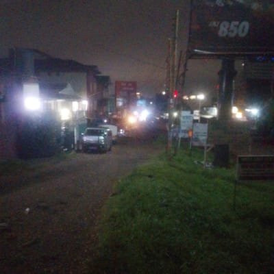 The image depicts a night scene in Ndenderu ward, Kenya. A dimly lit street leads towards a cluster of buildings and vehicles. A building with bright lights illuminating the surrounding area is on the left side of the street. Two cars, one white and the other silver, are parked along the street. A billboard with the number "850" is visible on the right side of the street. The foreground consists of a dark, grassy area. In the distance, there are street lights and the glow of other vehicles' headlights. A red traffic light can be seen in the mid-ground. Several small signs stand along the side of the road.