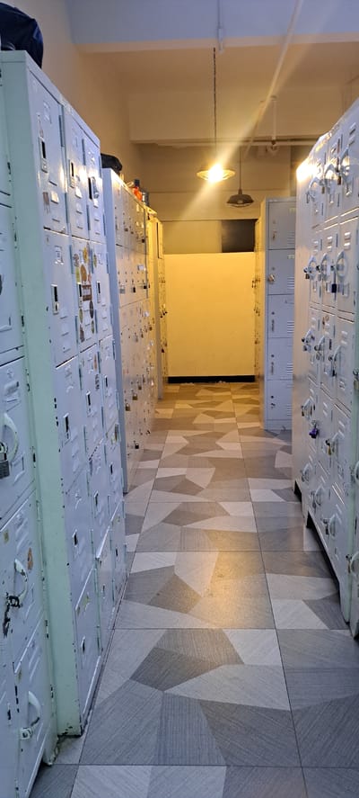 The image shows a hallway lined with rows of light-colored metal lockers on both sides. The lockers appear old and have various stickers and some padlocks attached to them. The hallway has a patterned tiled floor, with tiles in shades of gray, white, and beige arranged in a geometric design. Overhead, there are exposed pipes and two industrial-style pendant lights hanging from the ceiling, one of which is illuminated. The hallway extends into the distance, where it appears to open into a brighter, less defined space with a yellow wall. There are no people visible in the image. The scene suggests a utilitarian space, possibly a locker room or storage area, within a building.