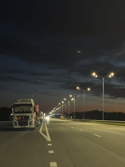 The image depicts a highway at dusk with a line of parked trucks on the left side of the road. The sky is dark blue with some clouds. Streetlights are illuminating the highway. The trucks appear to be stopped, possibly due to an overnight rest or traffic. The environment suggests a rural or semi-rural setting, as indicated by the dark trees lining the horizon. The image is taken at ground level, facing forward along the highway. No people are visible in the image. There is a small, bright green object in the upper right portion of the sky, the nature of which is unclear. The location is not specified as Alexeyevskoye, Russia.