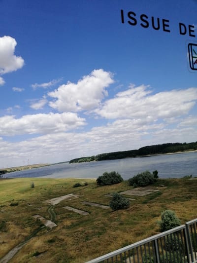 The image captures a scenic view of the Danube River near the village of Stelnica, Romania, on a bright, cloudy day. The foreground features a metal railing overlooking a grassy embankment dotted with patches of concrete foundations and overgrown vegetation. The middle ground showcases the wide, calm expanse of the Danube River, with a distant forested shoreline on the right and a power line pylons on the left bank. The sky is a vibrant blue, adorned with fluffy white clouds. In the upper right corner, a portion of a sign with the partial text "ISSUE DE" and a stylized symbol is visible. The overall impression is one of natural beauty and industrial remnants.