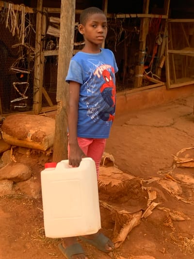 A young boy in a blue Spiderman shirt and pink pants stands near a wooden structure and red soil. He holds a white rectangular container with a red cap. The ground is uneven and covered with loose soil and stones. He wears blue sandals. The background includes a weathered structure with wire mesh.