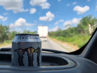 The image shows a close-up of a "Monster Energy Zero Sugar" can sitting in a car's cup holder. The can is silver with the distinctive black Monster Energy claw logo. The text "ZERO SUGAR" is clearly visible on the can.
The scene is viewed from inside the car, looking forward through the windshield. The background is blurred, suggesting motion. It appears to be a sunny day with a bright blue sky filled with fluffy white clouds. A road stretches ahead, and in the distance, a white truck is visible. The sides of the road are lined with green trees and foliage, typical of a rural or semi-rural landscape. The overall impression is one of travel, perhaps on a road trip or a journey through the Bulgarian countryside, given the location context of Totleben. There are no people visible in the image. The lighting suggests it is daytime, likely late morning or early afternoon.
