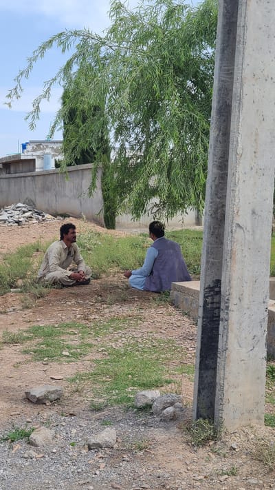 An outdoor scene in Qilla Saifullah, Pakistan, depicts two men resting on arid, rocky ground with patches of sparse green grass. The image is framed by a large, weathered concrete beam or structure running horizontally across the top.

In the foreground, a man with a dark beard and hair is seated on the ground, wearing a light-colored shalwar kameez. He is looking slightly upwards, with an open-mouthed expression, suggesting he might be speaking or listening intently. Further back and slightly elevated, another man is seated or crouched, facing away from the camera. He is dressed in a blue shalwar kameez and a dark blue or grey vest, seemingly looking down towards the first man or the ground in front of him.

To the left, a vibrant green tree with long, slender leaves, possibly a willow, dominates the mid-ground, its branches extending towards a clear, bright sky. In the background, beyond the men, several concrete walls and partial structures are visible, indicating an area under development or a simple settlement. A light blue water tank can be seen atop one of these structures. The ground is dry earth mixed with gravel and scattered rocks. The bright, clear sky suggests it is daytime, likely morning or afternoon, under dry weather conditions. No discernible text is visible in the image.