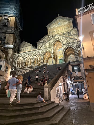 A nighttime exterior scene captures a grand cathedral with an ornate facade and a large set of stairs leading up to its entrance. The cathedral's architecture features striped arches and intricate geometric patterns. Several people are seated on the stairs or walking up them, some in pairs and some in small groups. A couple stands at the bottom of the stairs, the man with his arm around the woman. On the right side of the image, a narrow street runs alongside the stairs, lined with buildings that appear to be shops or restaurants. A streetlamp illuminates the scene. A man in a black apron stands on the street, and a motorcycle is parked nearby. Scaffolding is visible on a building to the left, suggesting ongoing construction or renovation. The image is taken from a low angle, looking up towards the cathedral. The location is identified as Pogerola, Italy.