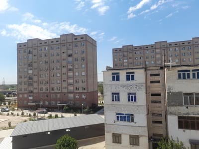 In Urgench, Uzbekistan, two multi-story apartment buildings are visible under a bright sky with scattered clouds. The building on the left is a larger, light-brown brick structure with numerous windows and balconies. In front of it, a covered parking area with a metal roof is situated, with a few cars parked nearby. To the right of the larger building, a smaller, weathered white apartment building with blue-tinted windows is partially obscured by the foreground. The environment appears to be a residential area with some sparse greenery and utility poles visible in the distance.