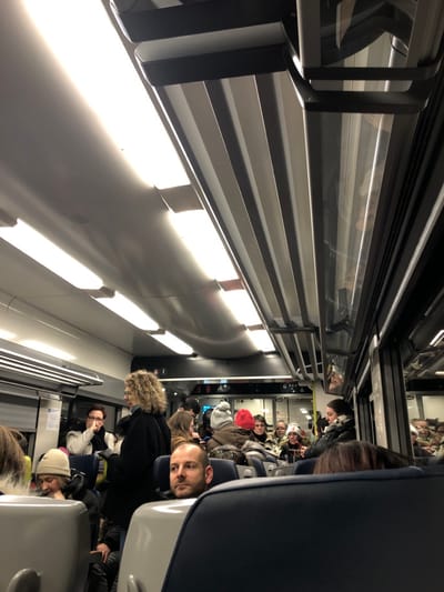 Passengers occupy seats within a crowded train car. The perspective is a long view down the aisle, showing the overhead luggage racks and the rows of seats filled with people. Some passengers are partially obscured, while others are more visible. The interior lighting is bright. A window runs along the top of the image. The seats are various colors.