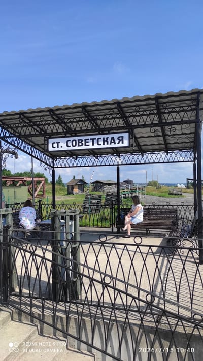 The image shows a covered outdoor waiting area at a train station. A sign above the platform reads "ст. СОВЕТСКАЯ". Two women are seated on a bench under the shelter. One woman on the left is wearing a white shirt with a colorful pattern and light-colored pants, and she is facing away from the camera. The woman on the right is wearing a white top and a light blue skirt, and she is looking to her left. Behind them, there are railroad tracks, some small buildings, and greenery under a blue sky with scattered clouds. A black metal fence runs in the foreground. The time stamp in the lower right corner indicates the image was taken on July 15, 2026, at 10:40 AM.