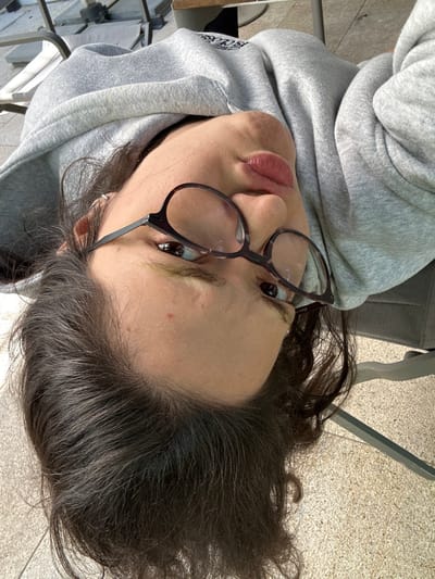 The image presents a close-up, inverted portrait of an individual situated outdoors in Vandœuvres, Switzerland, likely on a patio or terrace. The subject possesses dark, long hair and wears dark-framed eyeglasses, a light grey hooded sweatshirt displaying a partial dark emblem on the right chest, and a silver earring in the left earlobe. Their dark eyes are directed towards the camera, and their lips are slightly parted. A small red blemish is visible on the forehead. The background features light-colored outdoor paving, a silver-grey metal chair, and a light-colored flat surface, indicating an outdoor seating area under natural light.