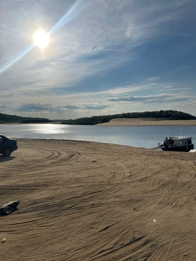 The image depicts a wide, calm body of water with a sandy shore in the foreground. A dark sedan is parked on the sand to the left, and a small boat with two people is positioned near the water's edge on the right. The sun is high in the sky, casting a bright glare on the water. The background shows a line of trees and sand dunes under a partly cloudy blue sky. Tire tracks are visible in the sand, indicating vehicle activity. The overall impression is a natural, somewhat remote riverside or lakeside scene, likely in a northern region given the vegetation and light. Pechora, Russia, is a city located in the Komi Republic on the banks of the Pechora River. This scene is consistent with the geography of the region, where rivers and sandy shores are common, and the climate can result in strong sunlight during summer months.