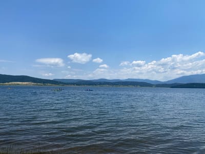 This is a wide-angle landscape photograph taken on a sunny day at Batak Reservoir in Bulgaria. The image captures a serene view of the expansive body of water, with gentle ripples on its surface reflecting the blue sky and scattered white clouds.
In the middle distance, several individuals are seen kayaking across the lake. There are at least three kayaks visible: one is yellow, another is lime green with two people in it, and a third is blue with two people. The kayaks are spaced out on the water, suggesting a leisurely activity.
The background of the image is dominated by rolling, tree-covered hills and mountains, which are layered and fade into the distance. The landscape suggests a natural, outdoor setting. The time of day appears to be late morning or early afternoon, given the bright sunlight and the shadows cast. The weather is clear and pleasant, with a bright blue sky and puffy white clouds. There are no visible signs of text in the image. The overall mood conveyed by the image is one of tranquility and outdoor recreation.