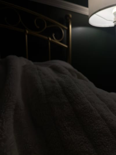 This is a close-up, low-light, eye-level shot of a plush, cream-colored blanket draped over a bed. The blanket has a textured, fluffy surface with visible folds and ripples, suggesting it is soft and warm. Behind the blanket, a portion of a gold-colored metal bed frame is visible, featuring decorative scrollwork and finials. To the right of the bed frame, a dark green wall is present, with a white lampshade casting a warm, soft light onto the wall and the top of the blanket. The lighting suggests it is nighttime or a dimly lit room. There is no discernible indication of the specific location within the City of Leduc, Canada, based solely on the visual information provided. The scene is static and focuses on the textures and lighting of a bedroom environment.